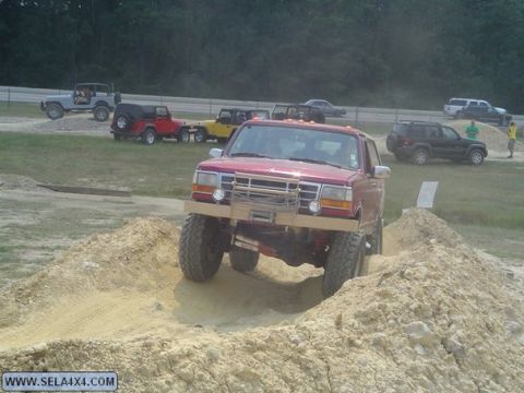 1995 Ford Bronco - A lifted red Ford truck drives over a dirt mound, with other off-road vehicles in the background.