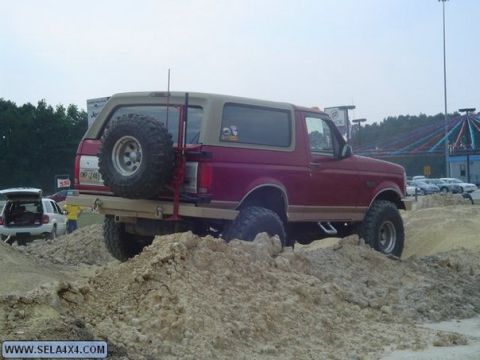 1995 Ford Bronco - A red and tan Ford Bronco navigates a dirt obstacle course with other vehicles in the background.
