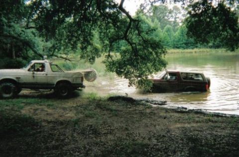 1995 Ford Bronco - Two full-size Ford Broncos at a muddy pond: a white lifted Bronco with a swing-out spare tire carrier sits on the bank while a dark red Bro…