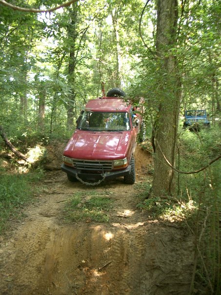 1995 Ford Bronco - A red SUV navigates a muddy trail through a dense forest, with another vehicle following behind.