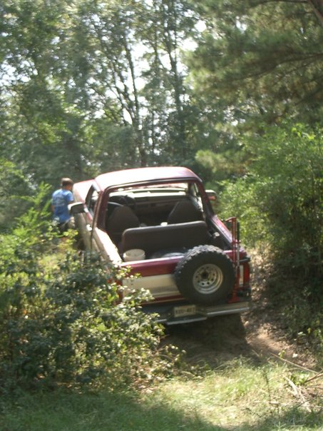1995 Ford Bronco - A red and white Ford Bronco is parked on a dirt path, partially obscured by foliage, with a person standing nearby.