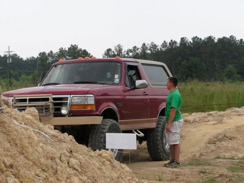 1995 Ford Bronco - A man stands beside a lifted maroon Ford Bronco with a tan top on a dirt track.