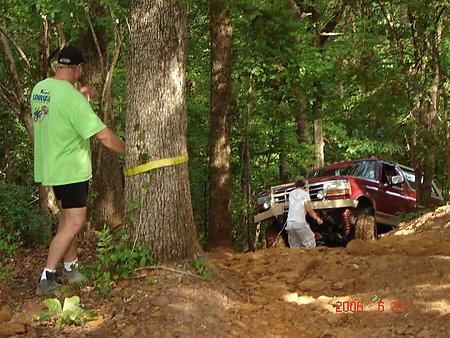 1995 Ford Bronco - A lifted red Ford Bronco sits nose-down in a dirt hole on a wooded trail while a man in a green shirt wraps a yellow tow strap around a tre…