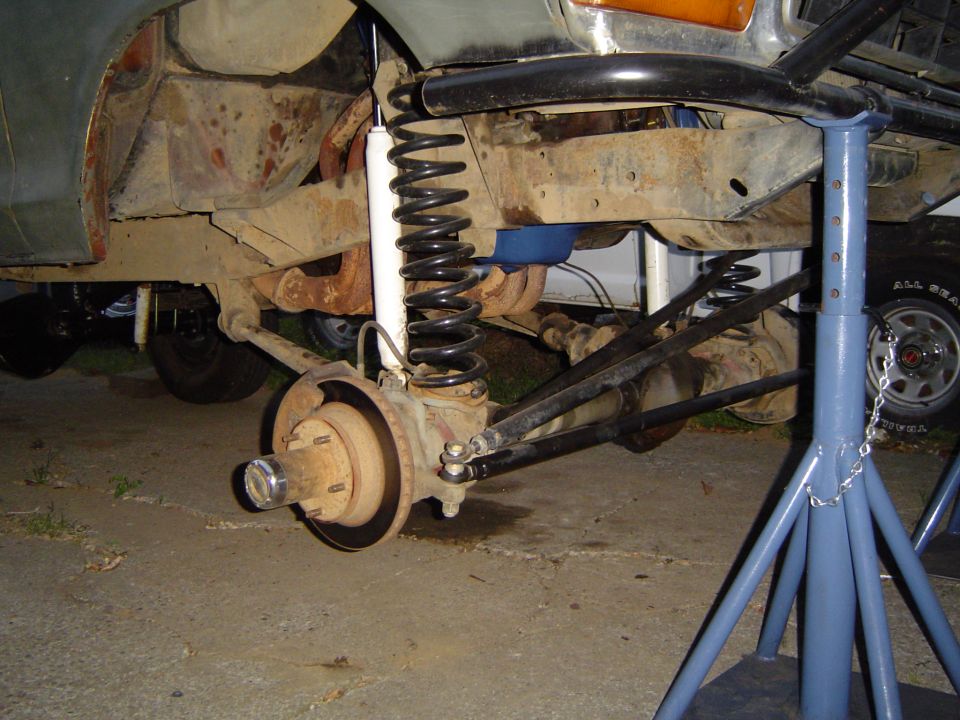 Front suspension and brake hub of a vehicle raised on a blue jack stand, showing coil spring and white shock.