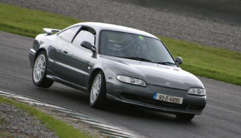 1991 Ford Probe - A gray Mazda MX-6 navigates a race track with its front-left wheel lifted, its sporty design and white racing wheels.