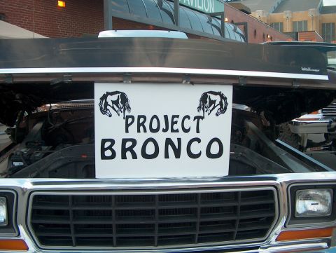 1978 Ford Bronco - The front grille and headlights of a classic truck are visible, with the hood raised to reveal the engine bay and a sign.