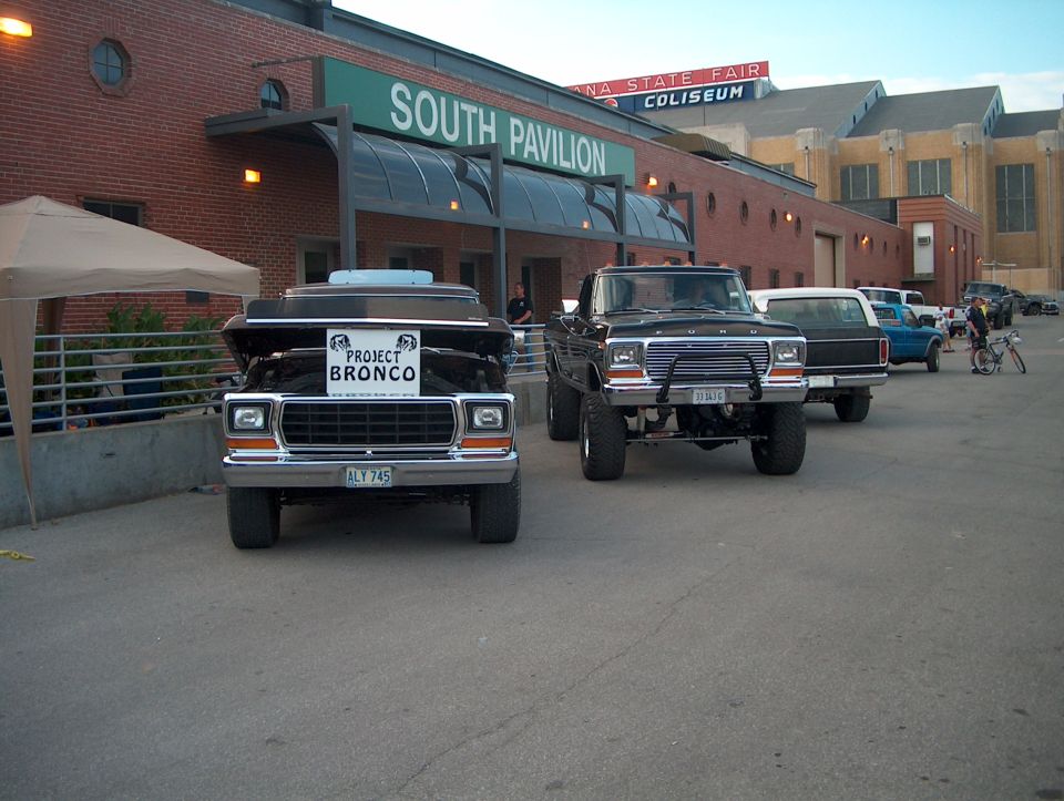 Front view of a black 1978 Ford Bronco with hood up and "PROJECT BRONCO" sign, parked next to a lifted black Ford pickup truck