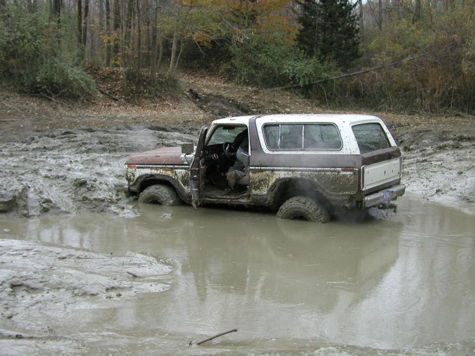 White and brown Ford Bronco SUV stuck deep in a muddy pit, driver door open