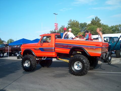 1978 Ford Bronco - Modified orange pickup truck with people in the bed, parked next to another truck.