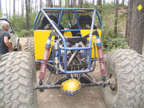 1989 Ford Bronco - A custom buggy with a yellow body and blue roll cage sits on a dirt path surrounded by trees.