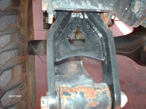 1982 Ford F-100 - Close underbody view of a boxed steel leaf-spring shackle bolted to a frame hanger, shot from below beside an aggressive mud-terrain tire w…