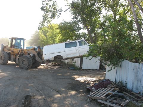 1988 Ford E250 - A white van is precariously balanced on a metal fence, being moved by a front-end loader.
