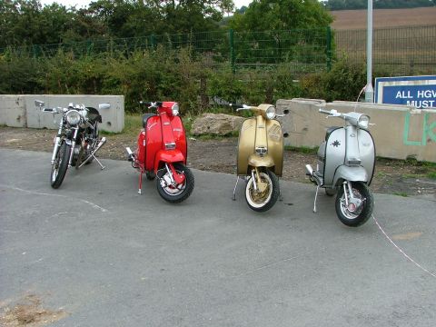 2001 Ford Fiesta - A motorcycle and three scooters, red, gold, and silver, are parked in a line on a paved area with a highway sign in the background.