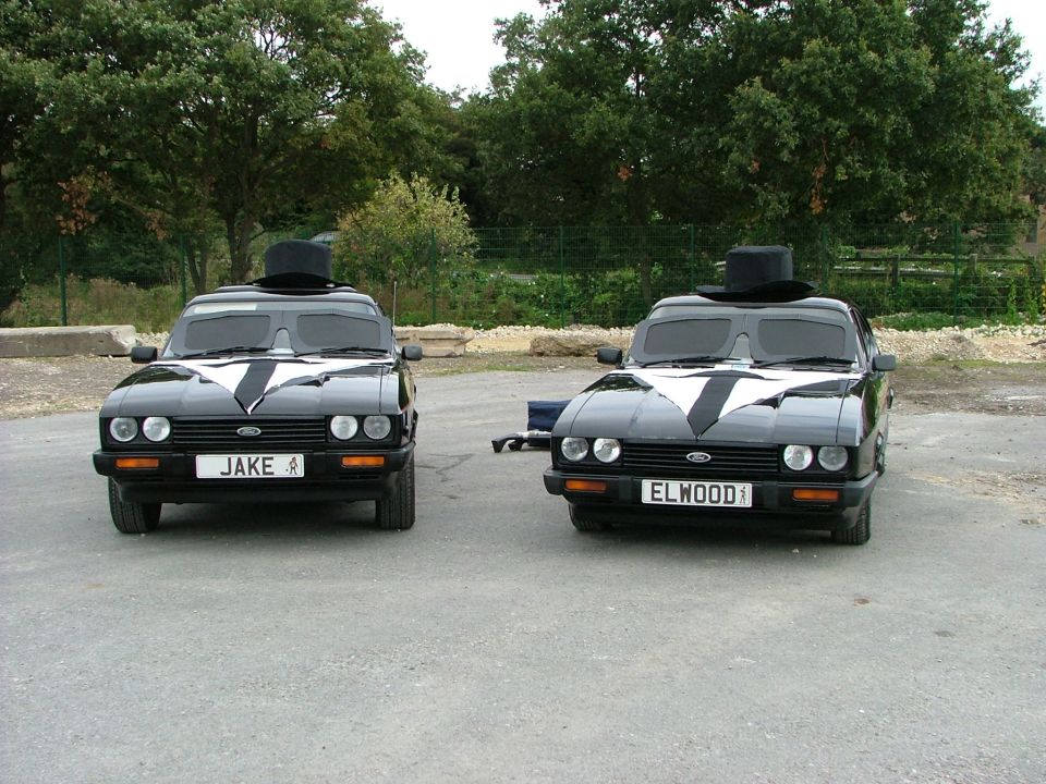 Two black Ford Capris with top hats and white tie decals, one with license plate JAKE, the other ELWOOD