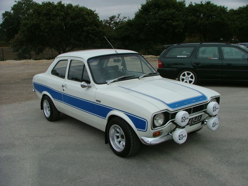 White 1970s Ford Escort RS2000 with blue stripes and rally lights. Green SUV in background