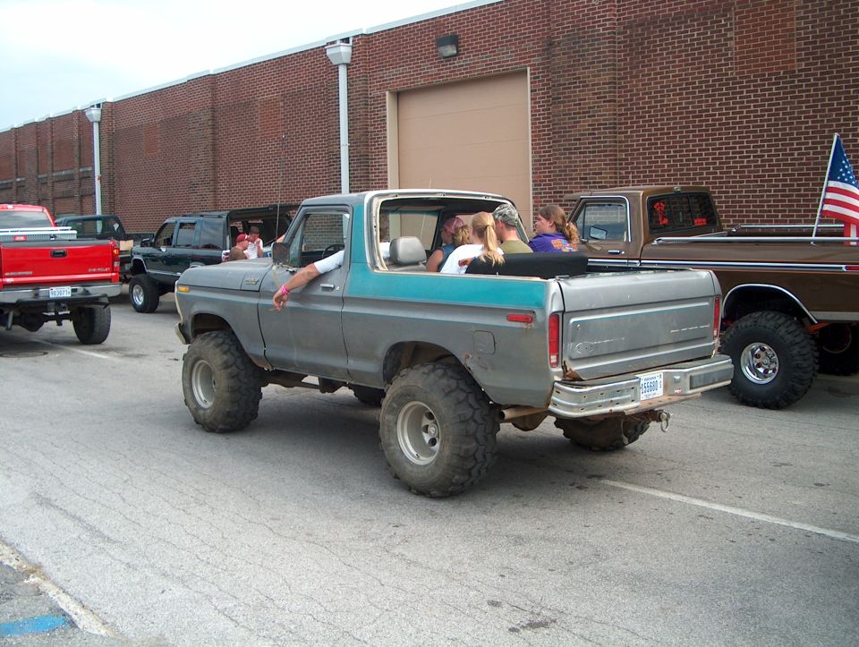 1978 Ford Bronco with lifted suspension and large tires, people in the cab. Another pickup truck is parked behind it