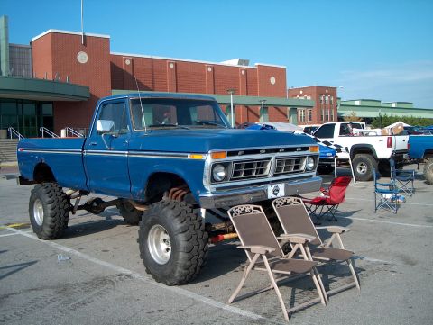 1978 Ford Bronco - A lifted blue pickup truck from the mid-1970s is parked in a lot with other vehicles and chairs.