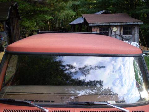 1979 Ford Bronco - Close-up of an orange SUV's roof and windshield, reflecting trees and clouds. A Supermotors logo is visible at the bottom.
