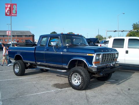 1978 Ford Bronco - A lifted blue Ford pickup truck is parked in a lot, with a white truck to its right.