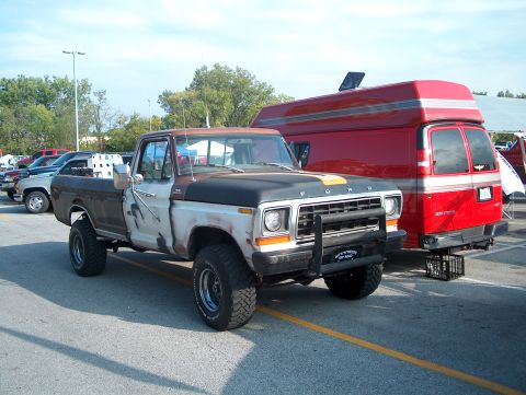 1978 Ford Bronco - A weathered 1978 Ford pickup truck is parked next to a red van, showing its front grille and lifted suspension.