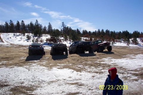 1978 Ford Bronco - Several off-road vehicles are parked on a snowy, dirt terrain with trees in the background.