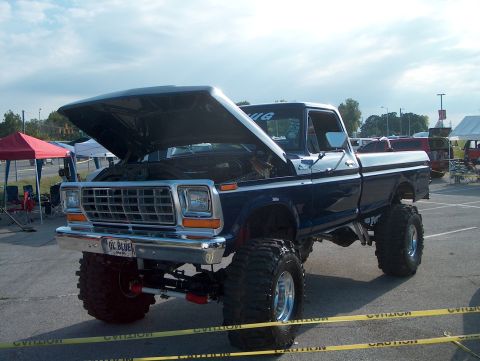 1978 Ford Bronco - Three-quarter front view of a lifted late-1970s Ford F-Series pickup in dark blue with a silver two-tone stripe, hood raised at a show behi…