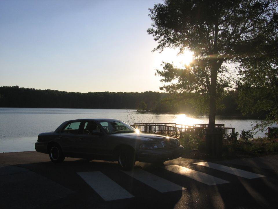 Dark sedan parked near a lake at sunset, with a tree and wooden dock in the background
