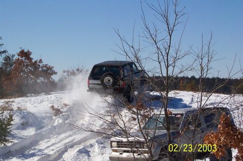 1978 Ford Bronco - Two SUVs navigate a snowy terrain, with one kicking up a spray of snow as it ascends a hill.