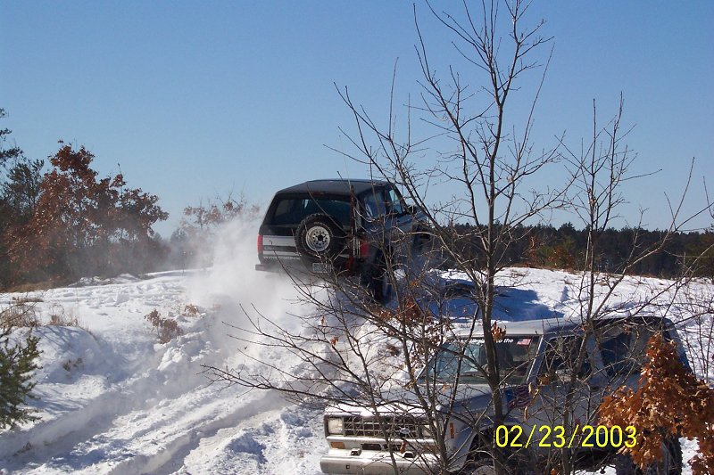 Two SUVs driving through snow. One is kicking up snow as it goes up a hill