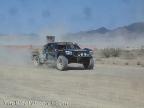 1990 Ford Bronco - black off-road race truck number 87 lettered Geiser Bros races through the dusty desert near a red fence.