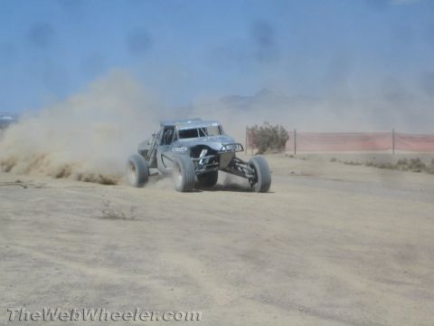 1990 Ford Bronco - gray single-seat off-road race buggy throws up a plume of dust as it powers along a desert dirt track.