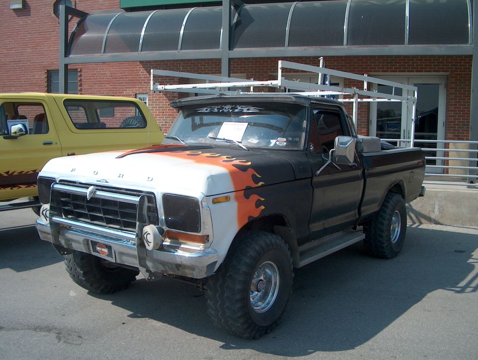 Black Ford pickup with orange and white flames on hood, and a ladder rack. Yellow Ford Bronco parked next to it