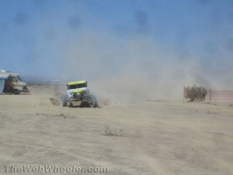 1990 Ford Bronco - yellow-and-white off-road race buggy speeds across a dusty open desert course past a parked support vehicle.