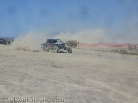 1990 Ford Bronco - blue off-road race buggy kicks up a tall dust cloud as it races along a desert dirt course lined with fencing.