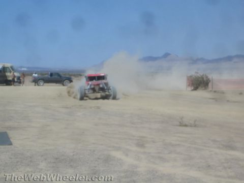 1990 Ford Bronco - red off-road race buggy raises dust on a desert flat as spectators and trucks watch from the sidelines.
