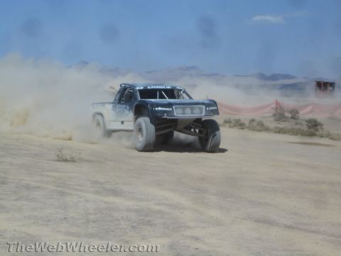 1990 Ford Bronco - dark off-road trophy-style truck throws up dust as it races across a desert course before distant mountains.
