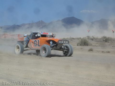 1990 Ford Bronco - orange open-cockpit off-road race buggy marked 01 races along a dusty desert course toward distant mountains.