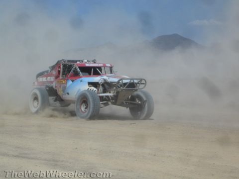 1990 Ford Bronco - red, white and blue off-road race buggy with sponsor decals races through heavy dust on a desert course.