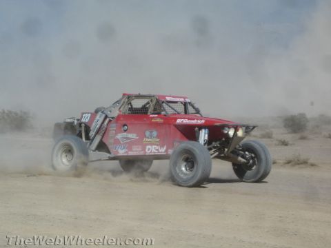 1990 Ford Bronco - red off-road race buggy covered in sponsor decals races along a dusty desert track lined with brush.