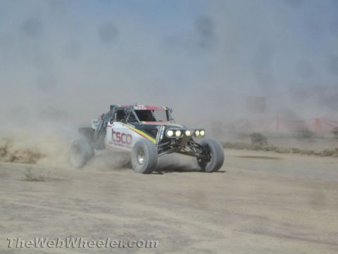 1990 Ford Bronco - black off-road race buggy with front light pods speeds through a thick dust cloud on a desert course.