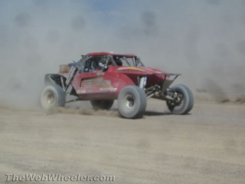 1990 Ford Bronco - red off-road race buggy viewed from behind kicks up dust as it accelerates along a desert track.