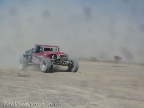 1990 Ford Bronco - pink and red off-road race buggy speeds across a dusty desert flat surrounded by haze.