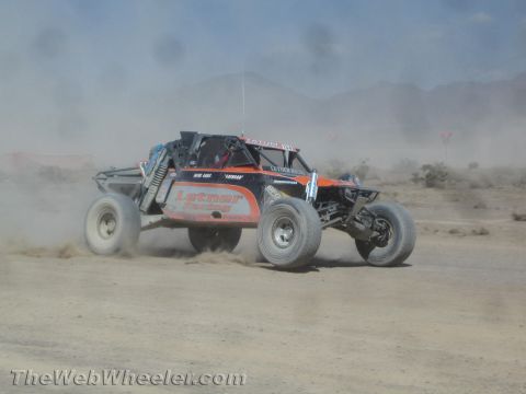 1990 Ford Bronco - orange and black off-road race buggy with sponsor graphics races along a dusty desert course below mountains.
