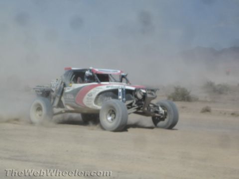 1990 Ford Bronco - red and white off-road race buggy kicks up dust as it races along a desert dirt course.