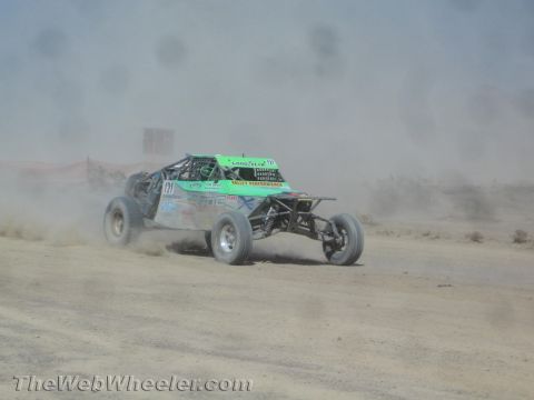 1990 Ford Bronco - green and white off-road race buggy with sponsor decals speeds through a dusty desert course.
