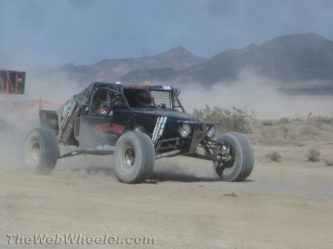 1990 Ford Bronco - black open-cockpit off-road race buggy speeds along a dusty desert course with mountains in the distance.
