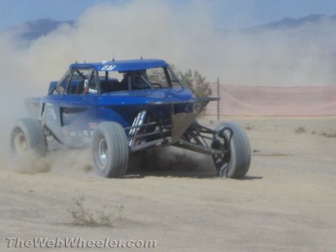 1990 Ford Bronco - blue off-road race buggy raises dust as it races across an open desert course lined with fencing.