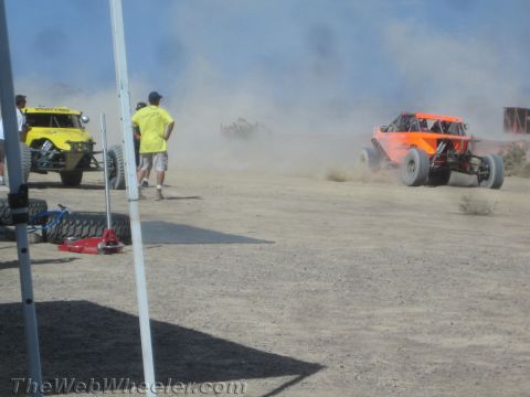1990 Ford Bronco - Off-road race buggies kick up dust on a desert course as spectators and a yellow buggy sit in a pit area in the foreground.