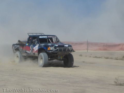 1990 Ford Bronco - dark off-road race truck marked 706 throws up dust as it speeds along a desert course past orange fencing.