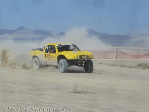 1990 Ford Bronco - yellow off-road race truck kicks up dust as it races across an open desert course toward distant mountains.